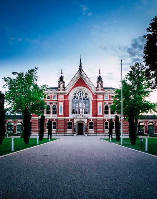 A large historic building with a red brick facade, ornate white detailing, and multiple arched windows is visible in the image. The structure features a central entrance with a decorative stone surrou