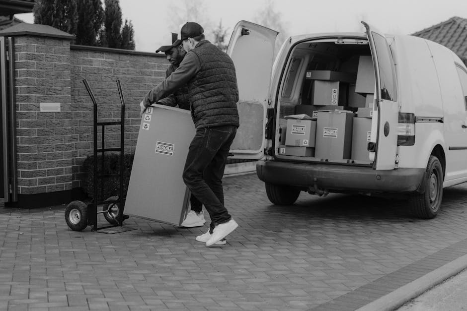A man wearing a blue jacket and dark trousers is standing outside a white moving van with open rear doors on a cobblestone street in East Dulwich. He is holding a cardboard box, preparing for a home r