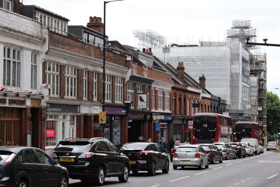 A busy street scene in East Dulwich showing a row of terraced houses with brick facades and white window frames, some of which are under scaffolding and construction work. Several vehicles, including